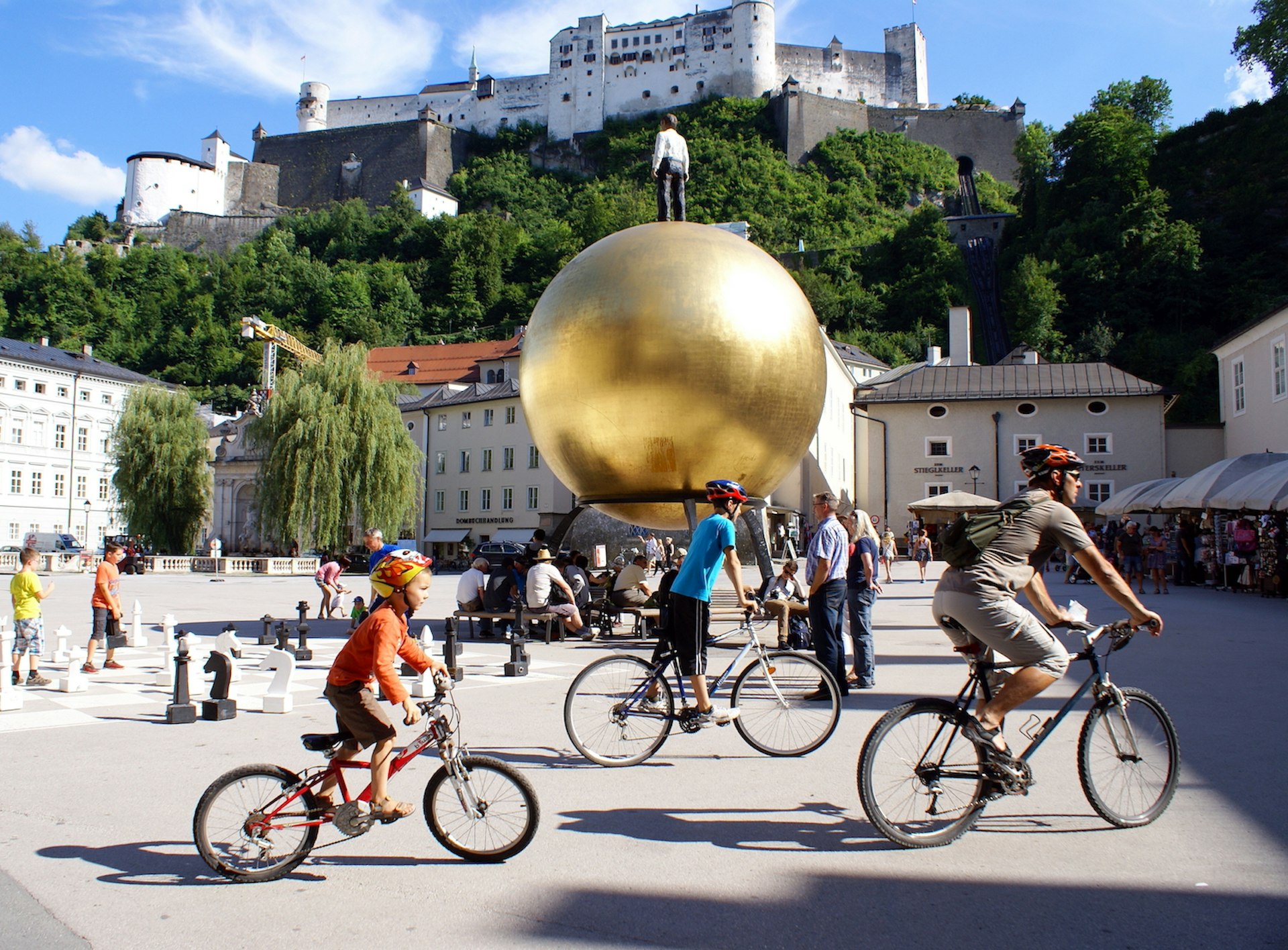 Cyclists pedal past the “Sphaera” sculpture and Hohensalzburg Fortress in the distance in historic Salzburg, Austria, Europe
