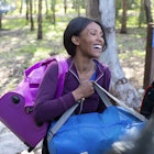 A happy woman traveling, loading purple and blue bags into the car trunk. Close up smiling young woman packing / unpacking luggage from the car trunk. View from outside of the car.