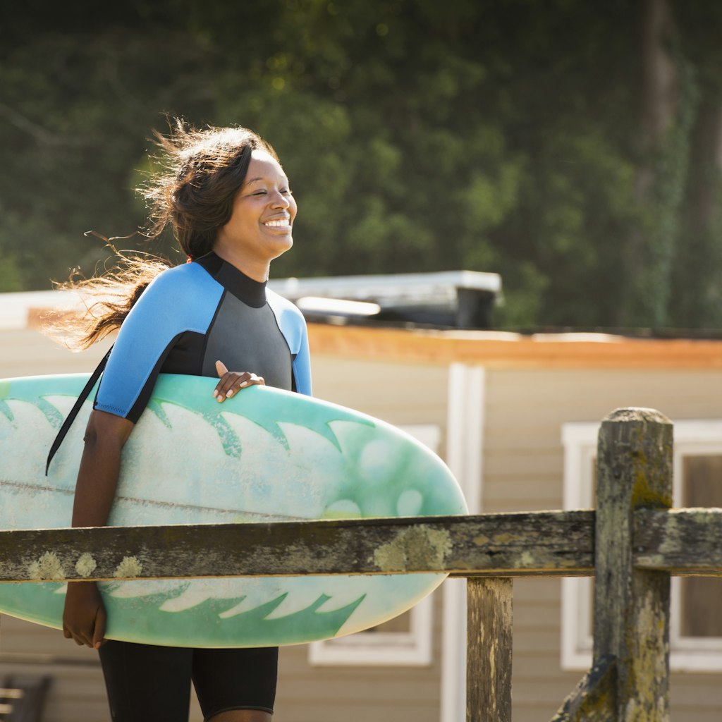 Smiling African-American woman walks down a pier in a wetsuit carrying a surfboard