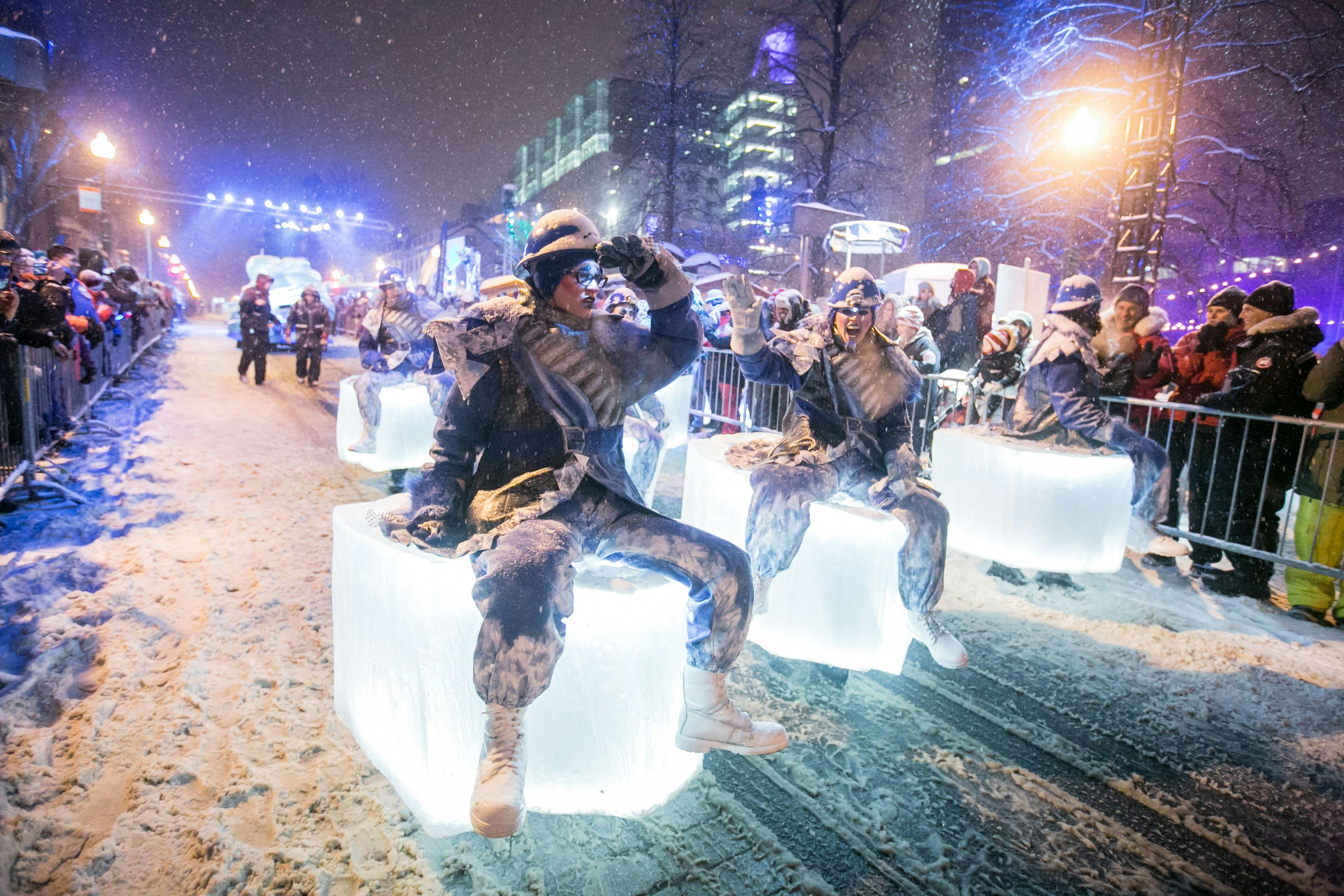 People in winter clothing in a parade, sitting on top of glowing cubes with snow on the ground and spectators lining the street
