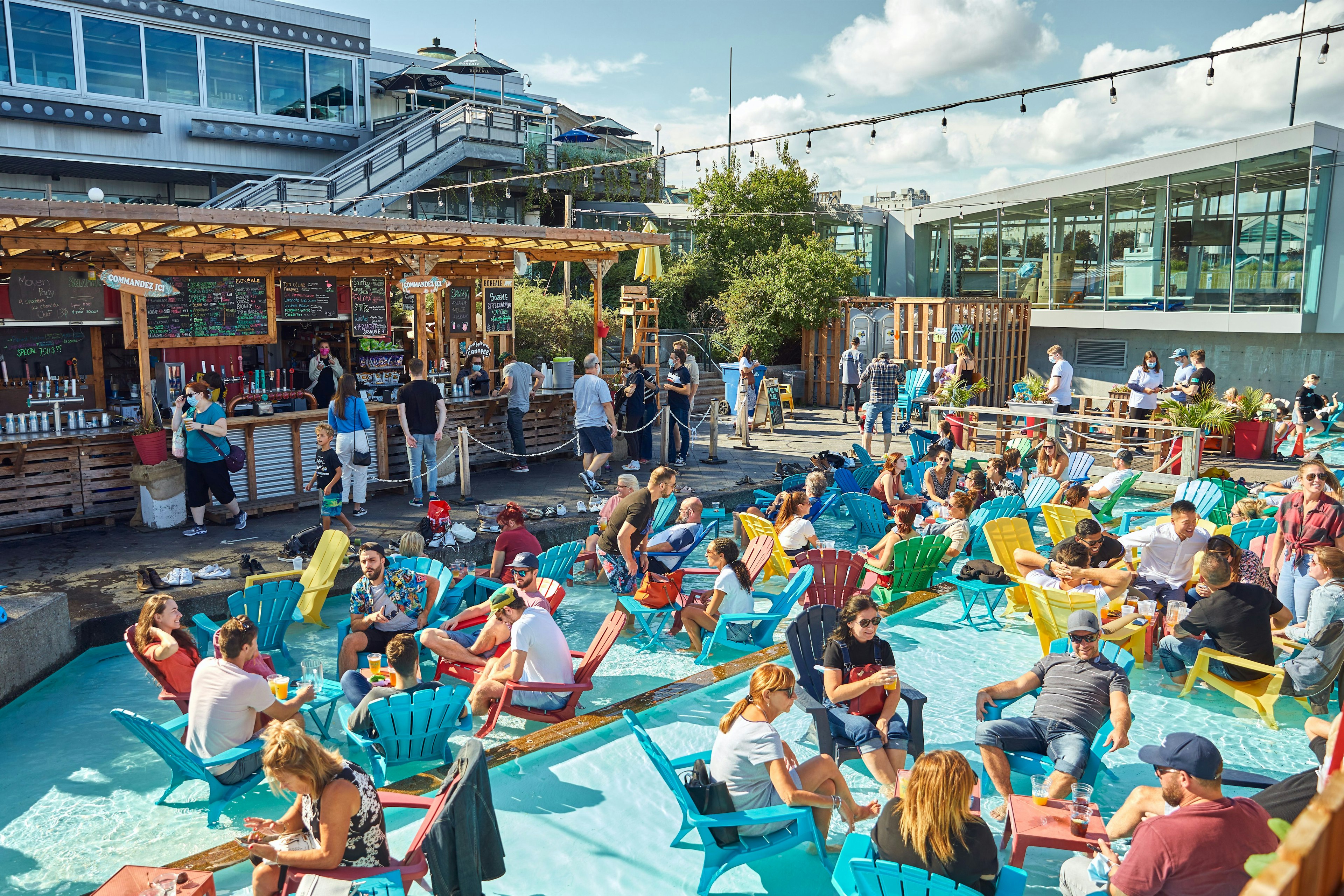 A crowd of people sitting in a shallow pool in adirondack chairs, with a bar in the background