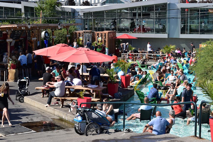 A crowd of people sit at picnic tables and chairs in the wading pools at la Cour arrière du Festibière, Québec City, Québec, Canada