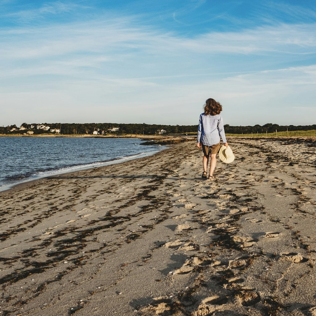 A woman holding a straw hat walks along the shore on a beach in Cape Cod, Massachusetts.  