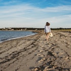 A woman holding a straw hat walks along the shore on a beach in Cape Cod, Massachusetts.  