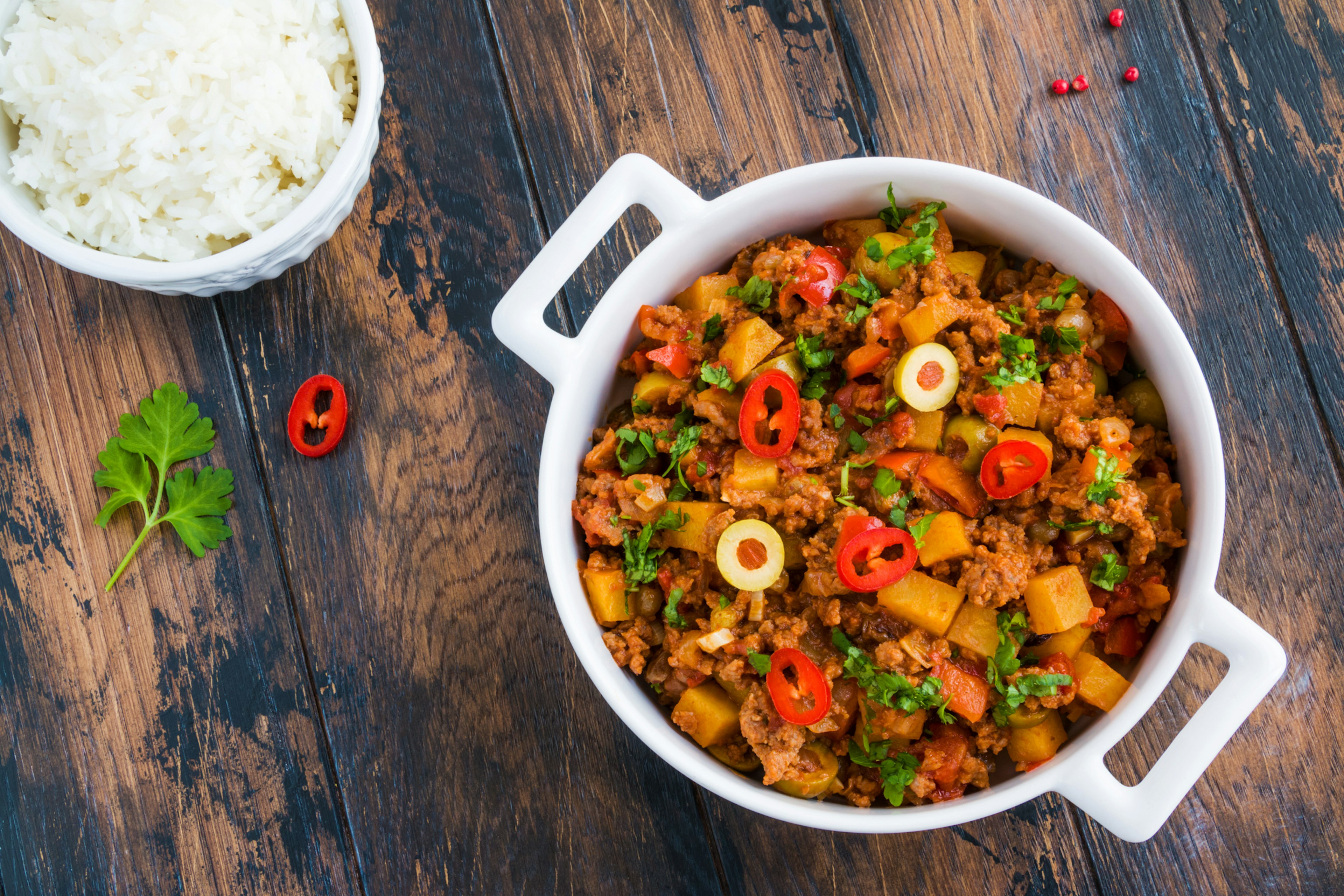 Classic Cuban dish picadillo a la habanera, a soft, fragrant stew of ground beef and tomatoes, with raisins and olives, in a white casserole and boiled rice on the wooden rustic table, top view.