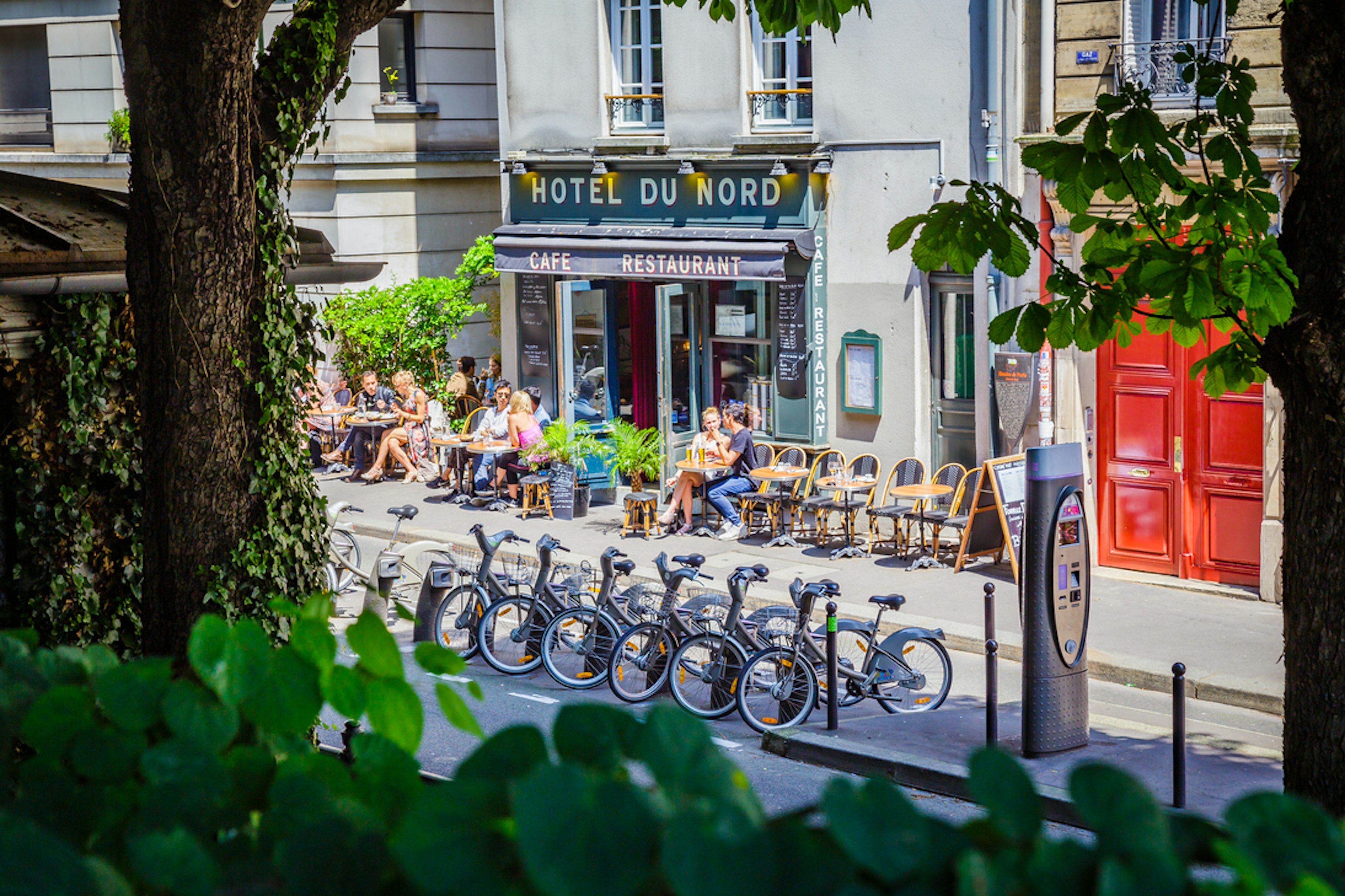 People sat at tables outside a cafe called Hotel du Nord. The street is lined with bicycles