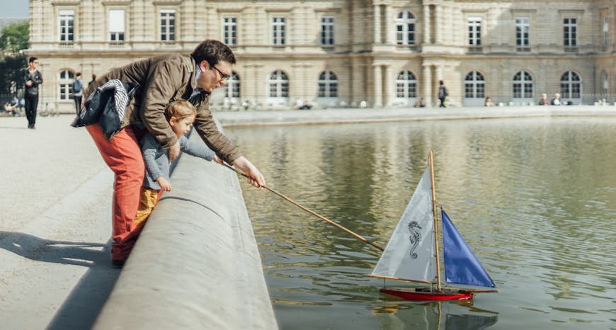 A father and son push a small sail boat out onto a pond in front of a palace