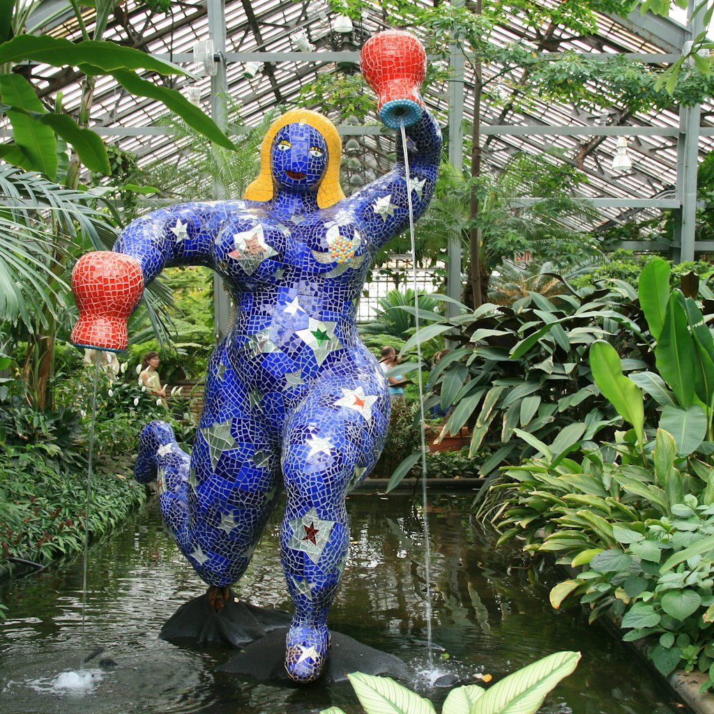 A colorful blue statue with bright yellow hair in the shape of a woman holding two orange pots with water pouring out of them. The statue is surrounded by greenery inside the Garfield Park Conservatory.  