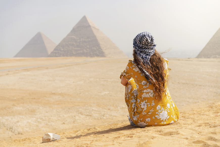 A female tourist sitting on a sand dune and looking at the Pyramids of Giza. 