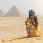 A female tourist sitting on a sand dune and looking at the Pyramids of Giza.