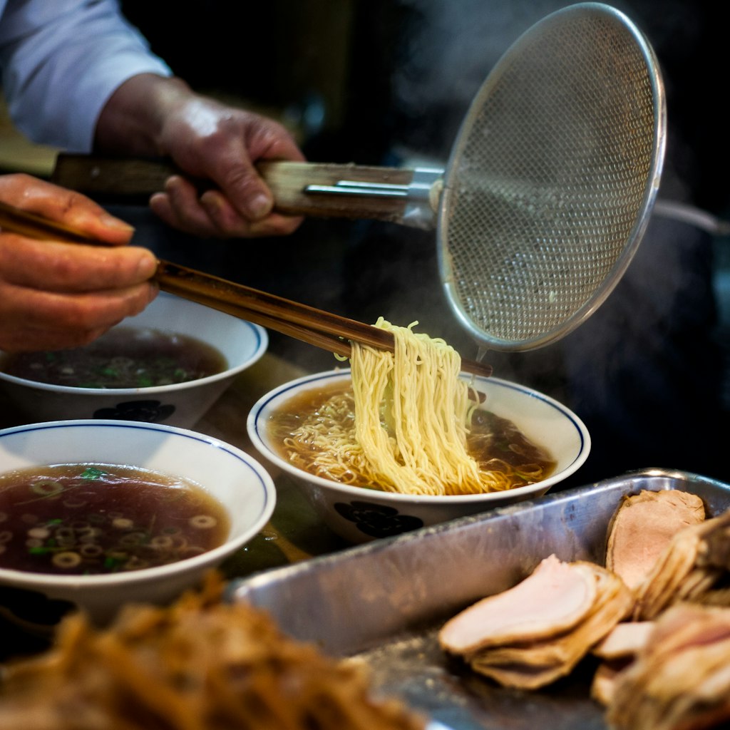 Hands preparing steaming ramen (soba) while holding long chopsticks and metal strainer. Several bowls of broth and pork are in the foreground.