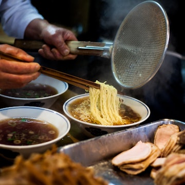 Hands preparing steaming ramen (soba) while holding long chopsticks and metal strainer. Several bowls of broth and pork are in the foreground.