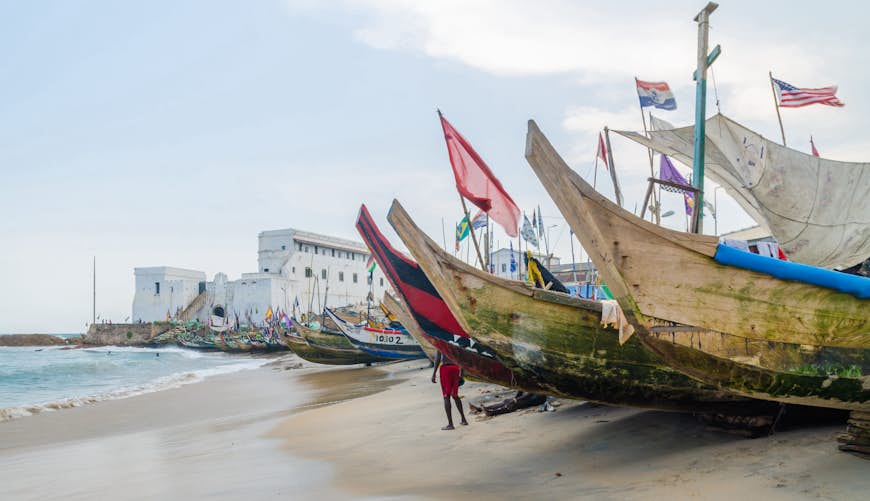 Färgglada förtöjda träfiskebåtar kantar stranden med slavslottet i bakgrunden, Cape Coast, Ghana, Västafrika