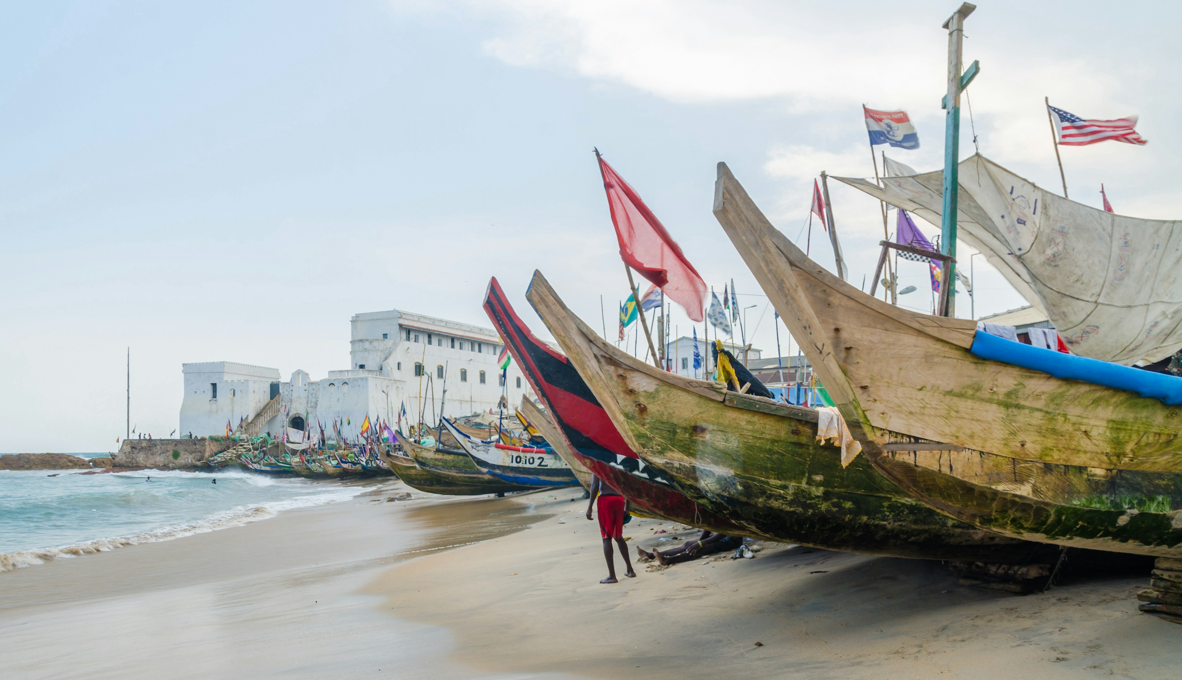 Colorful moored wooden fishing boats line the beach with the slave castle in the background, Cape Coast, Ghana, West Africa