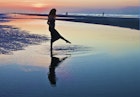 A woman in silhouette wearing a long dress  raises her foot out of the water during low tide at  sunset.  