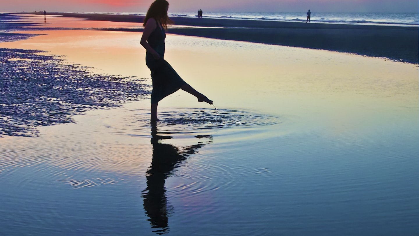 A woman in silhouette wearing a long dress  raises her foot out of the water during low tide at  sunset.  