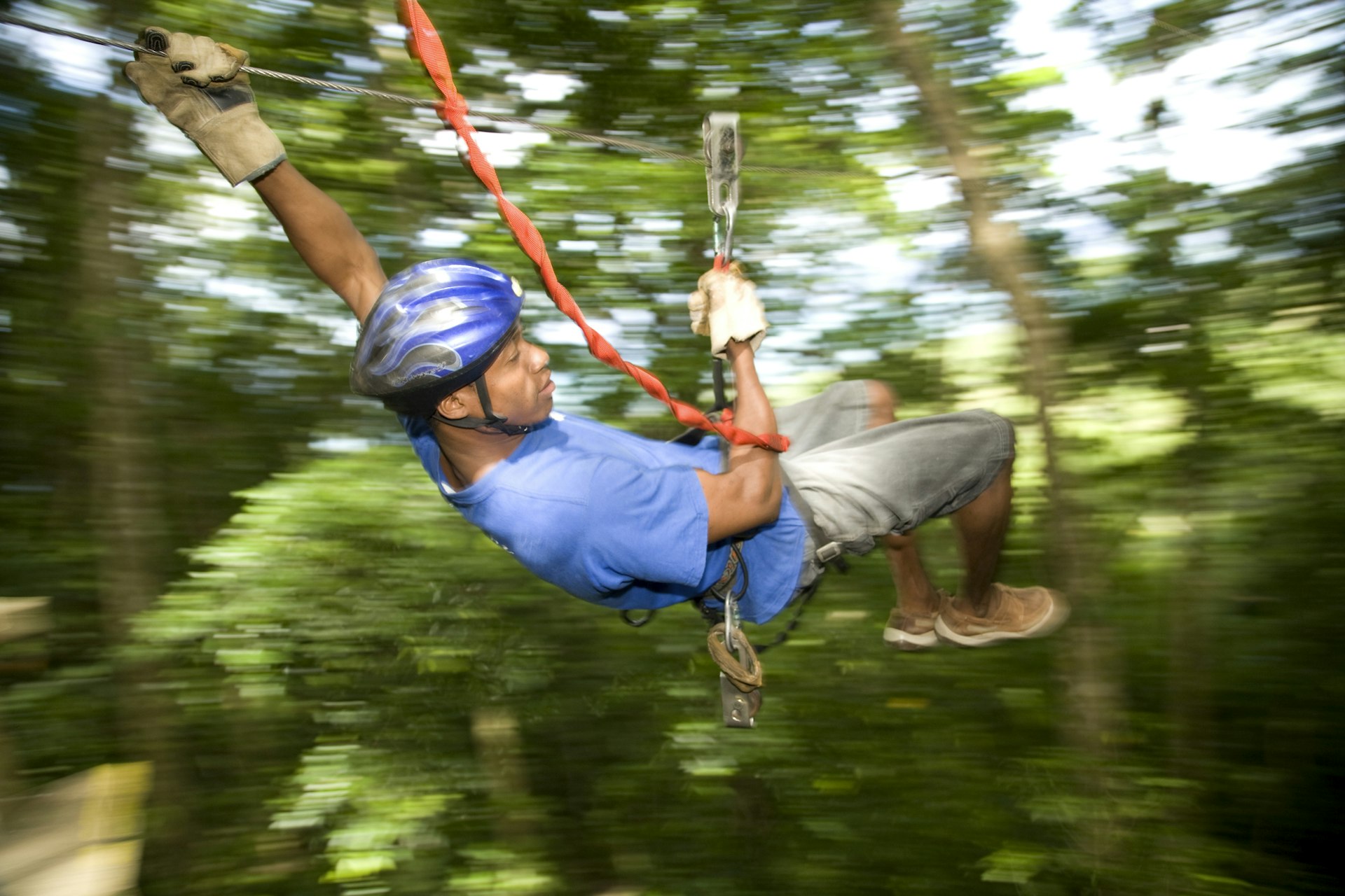 A Black man in a blue shirt, shorts and blue helmet ziplines through the Honduran rainforest