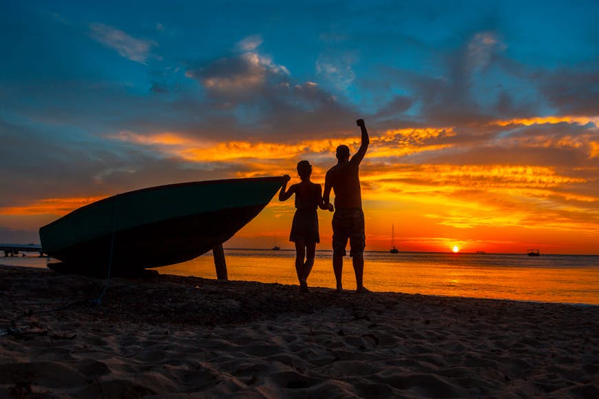 Una pareja al lado de un bote está recortada por la brillante luz roja y naranja de una puesta de sol, Roatán, Honduras, América Central