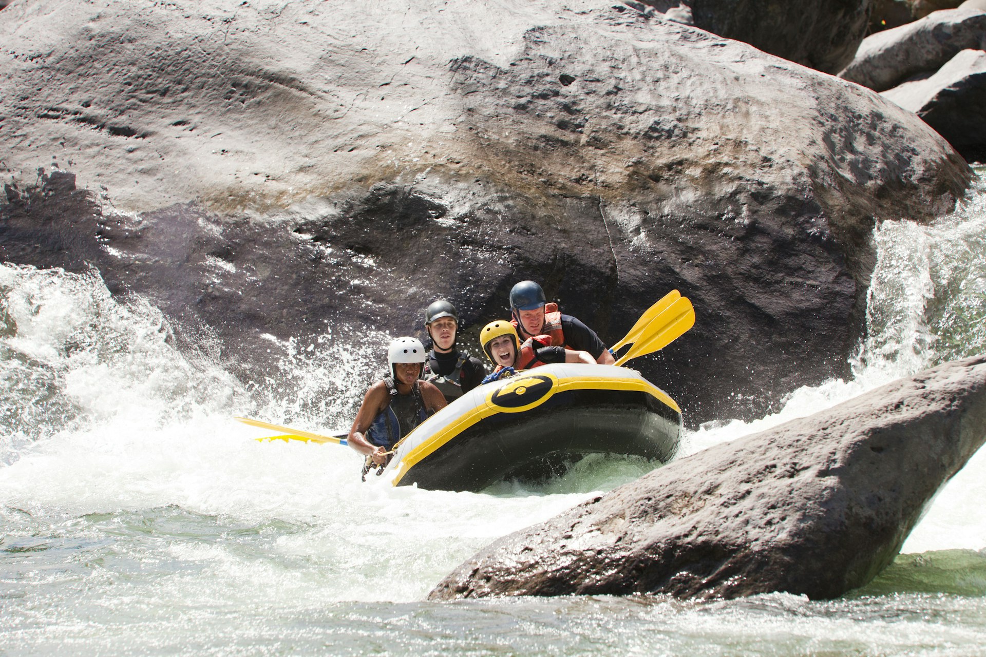 A group of four people, all wearing helmets, sit aboard a white-water raft as they hurtle down the Río Cangrejal in Honduras with huge boulders in the background