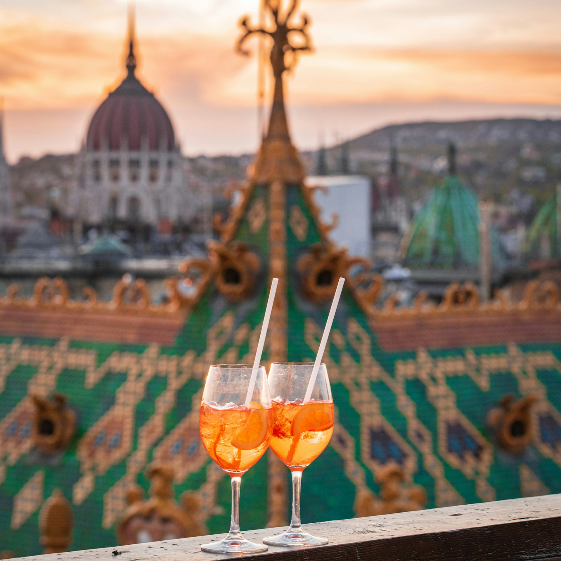 A pair of cocktails at sunset on a rooftop ledge in focus, with buildings including the Parliament in the background, Budapest, Hungary, Europe