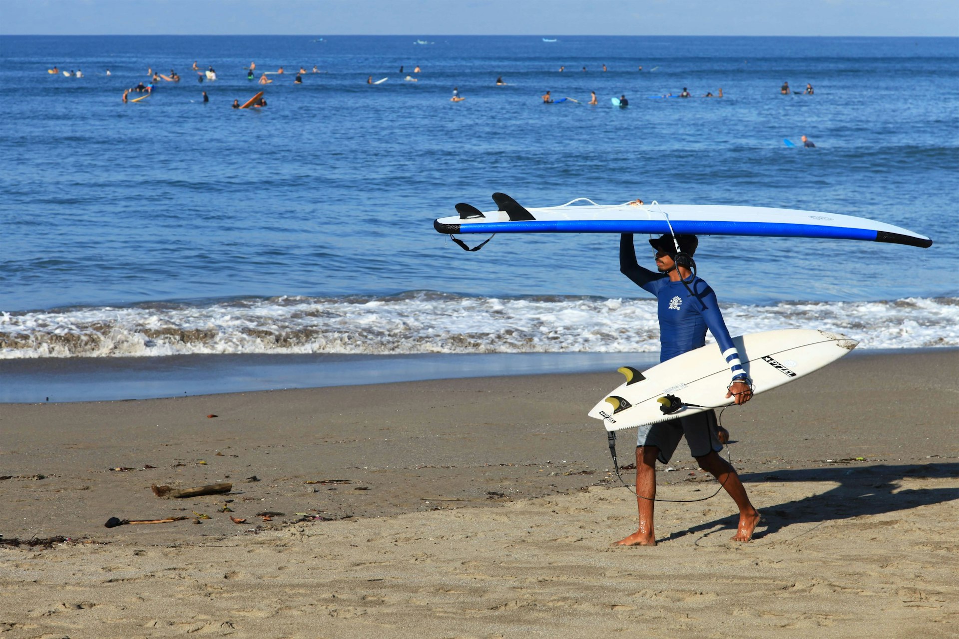 An Indonesian surf instructor walking on the beach carrying a two surfboards with one on his head at Batu Bolong Beach in Bali and surfers in the sea.
