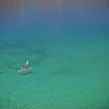 Three children on a paddle board one is standing the other two sitting. They are paddling on a vast turqoise sea, photo taken from above