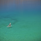 Three children on a paddle board one is standing the other two sitting. They are paddling on a vast turqoise sea, photo taken from above