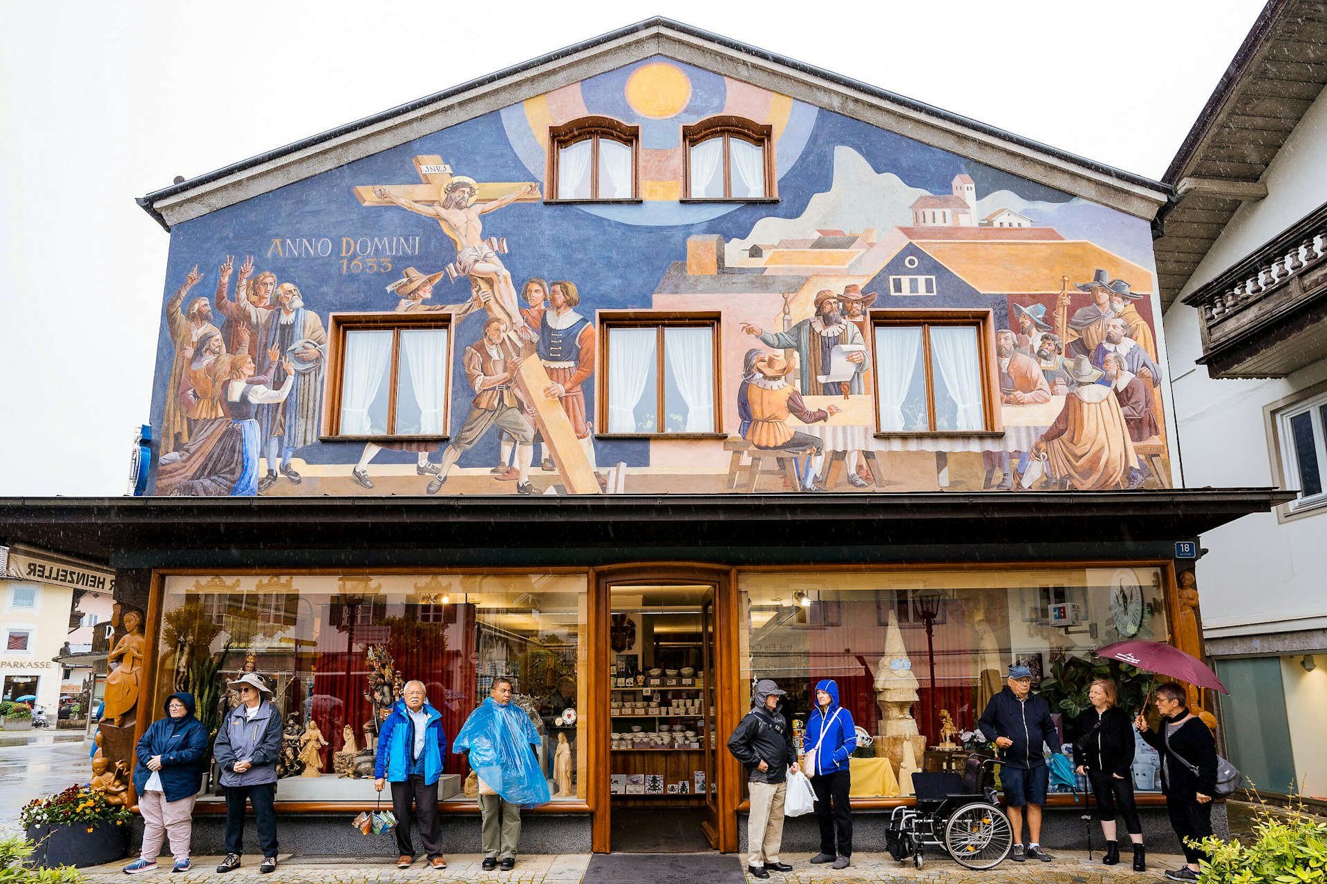 Tourists hide from the rain under the awning of a building with a painted fresco of the village's historic Passion Play 