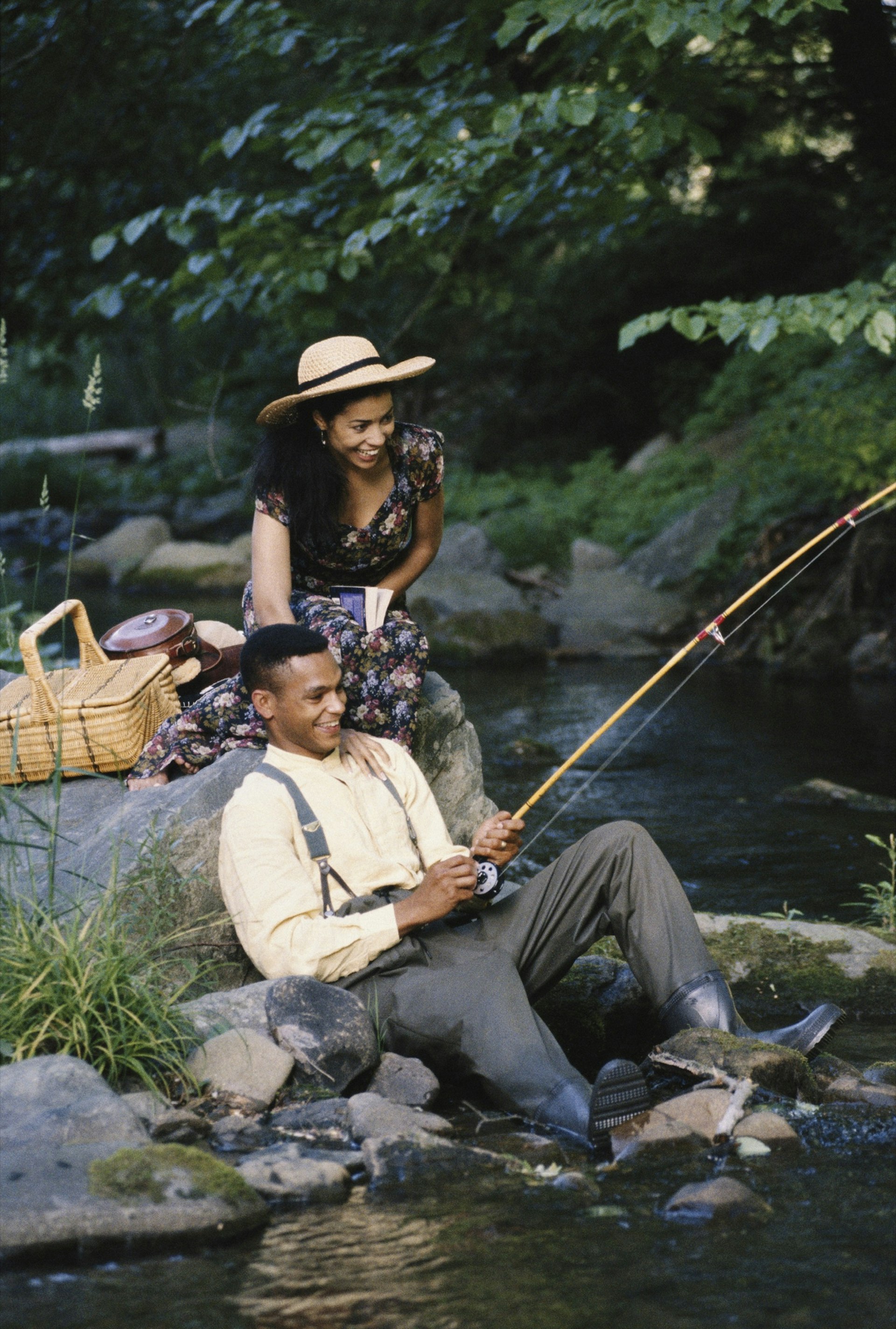 A man holds a fishing pole while a woman touches his shoulder and watches. There's a wicker picnic basket resting on the rocks.  