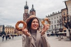 Tourist woman eating bagel obwarzanek traditional polish cuisine snack waling on Market square in Krakow. Traveling Europe in autumn; Shutterstock ID 2057885801; your: Claire Naylor; gl: 65050; netsuite: Online ed; full: Krakow budget