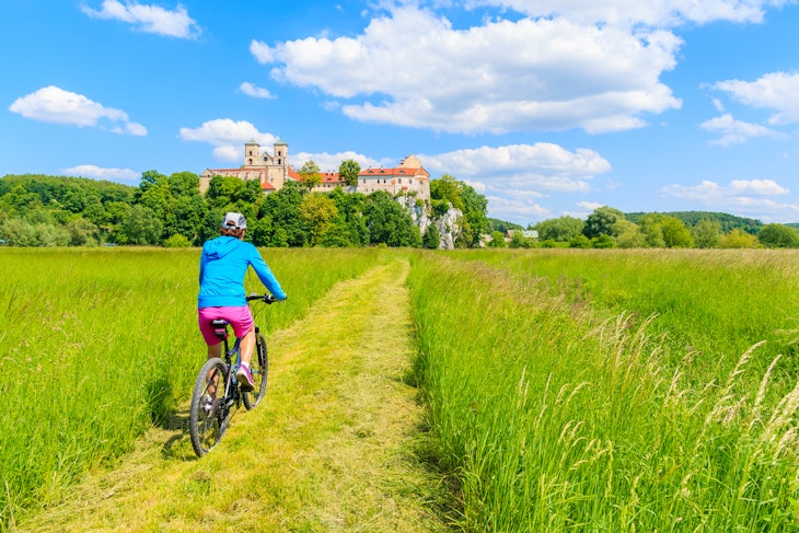 Young woman cyclist riding bike on green meadow and view of Tyniec monastery on sunny spring day, Cracow city, Poland

