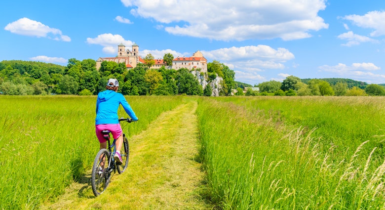 Young woman cyclist riding bike on green meadow and view of Tyniec monastery on sunny spring day, Cracow city, Poland

