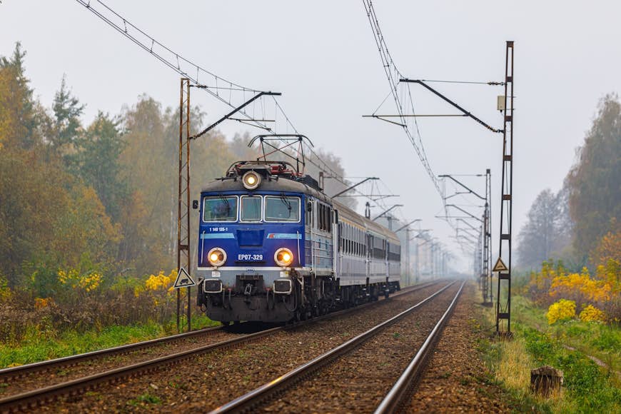 Blue EP07 locomotive pulls the carriages of the intercity passenger train to Warsaw through the autumn forest, Poland