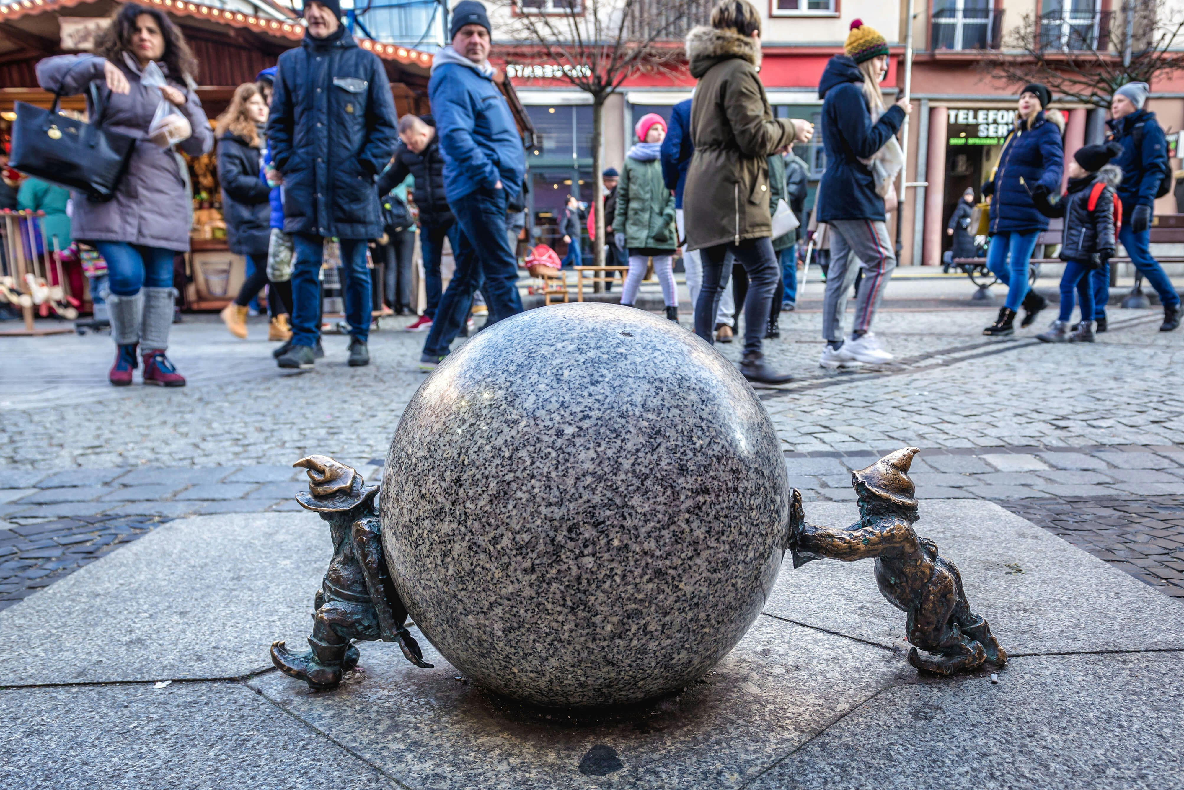 Two small dwarf statues on either side of a round marble ball, both pushing inward, with a tourist crowd looking at them in the background