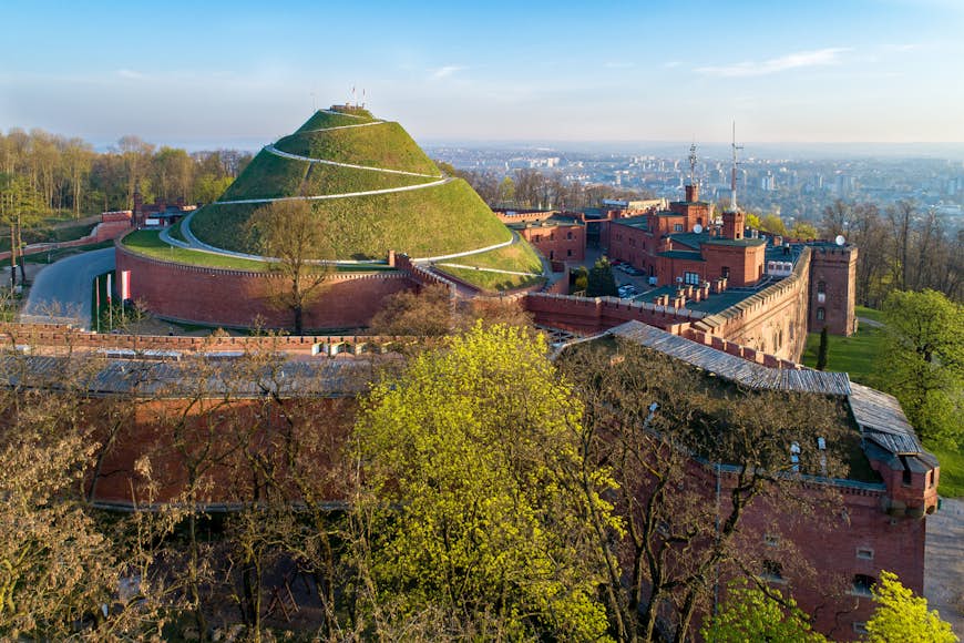 A huge artificial mound with a path circling it and a view of the city of Krakow in the distance