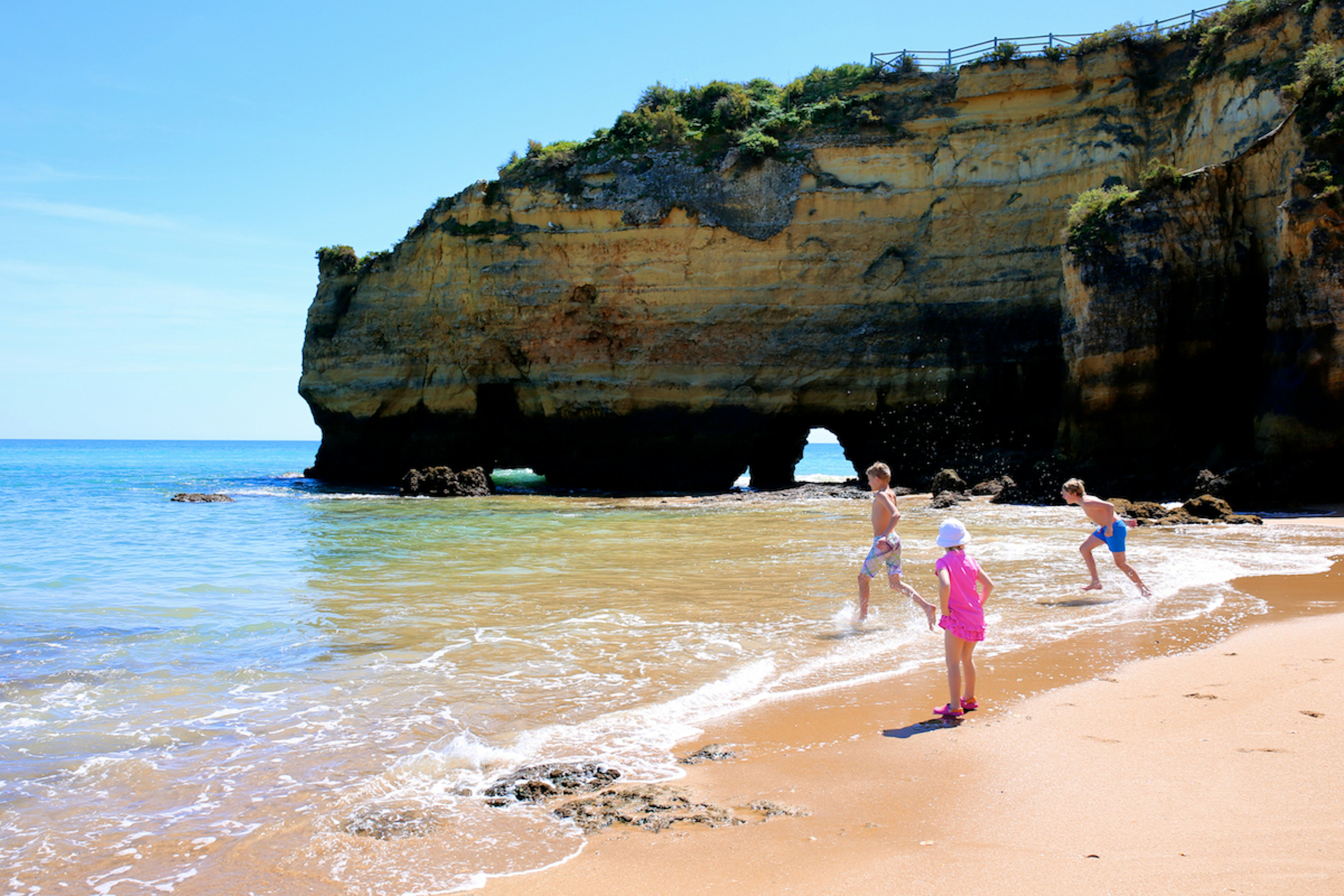 Happy children playing, splashing and swimming in the ocean against dramatic cliffs at Praia dos Estudantes, the Algarve, Portugal