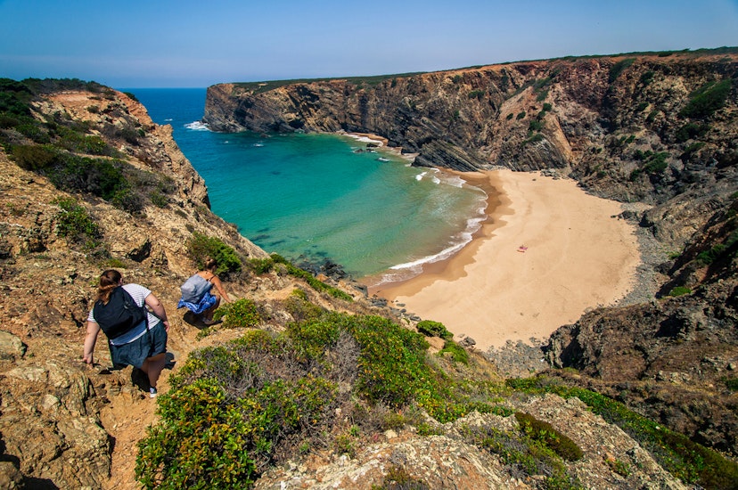 Landscape photo of two women climbing down a hill to a big, beautiful, nearly deserted beach with turquoise waters. Shot in Parque Natural do Sudoeste Alentejano e Costa Vicentina, Portugal. 