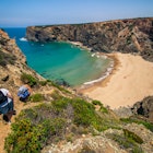 Landscape photo of two women climbing down a hill to a big, beautiful, nearly deserted beach with turquoise waters. Shot in Parque Natural do Sudoeste Alentejano e Costa Vicentina, Portugal.