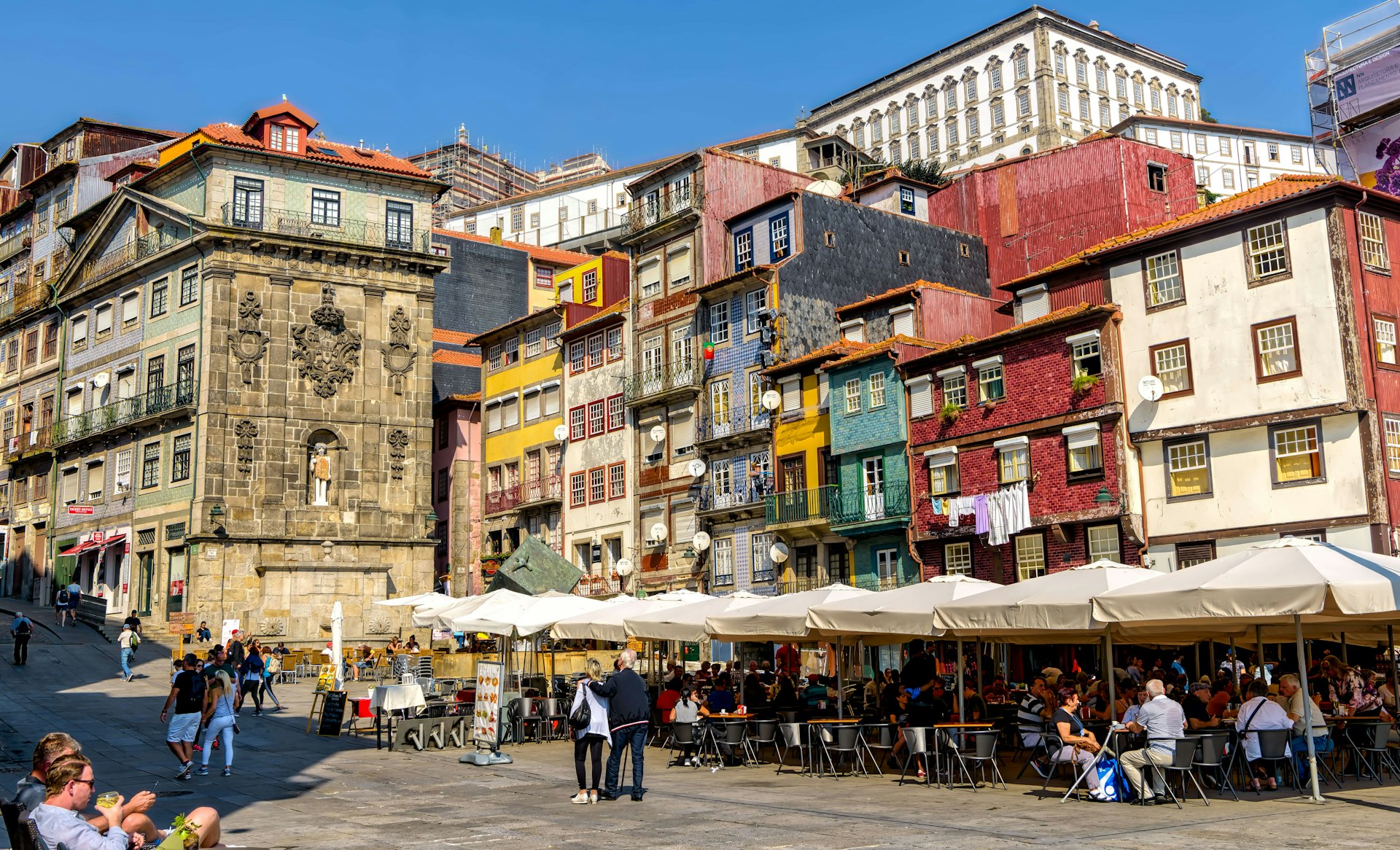 People walking and sitting at sidewalk tables in the Ribeira neighborhood, with a backdrop of tall, colorful buildings