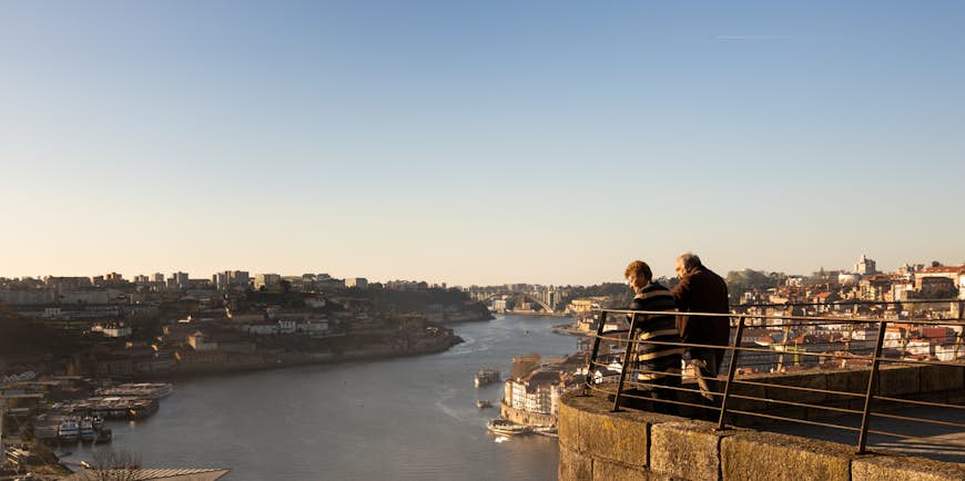 A senior couple stand together at the railings of a viewpoint looking down over a city with a river as the sun sets