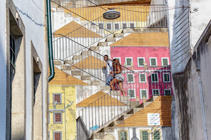 A couple walk up a set of stairs covered in street art depicting the sides of buildings and rooftops, creating an effect where it seems as though the couple is walking on roofs