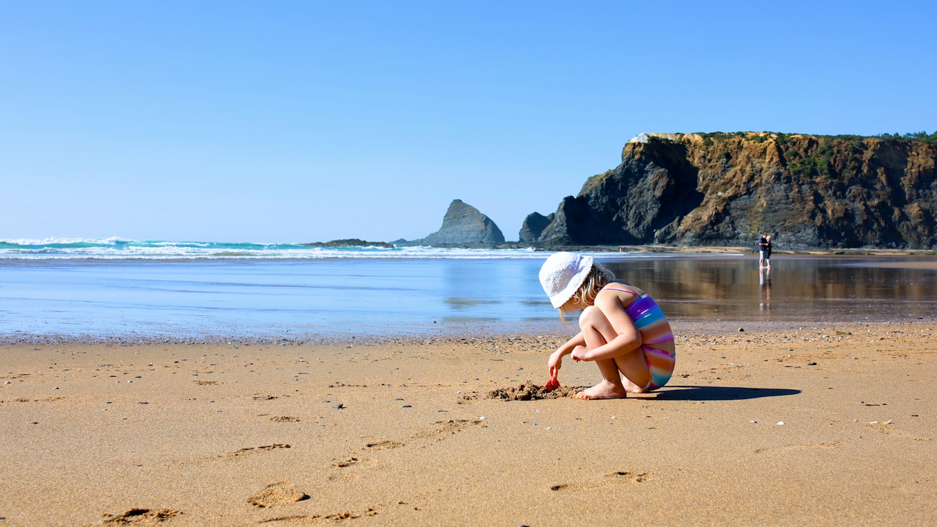 A young child plays with a spade on a sandy beach backed by large cliffs