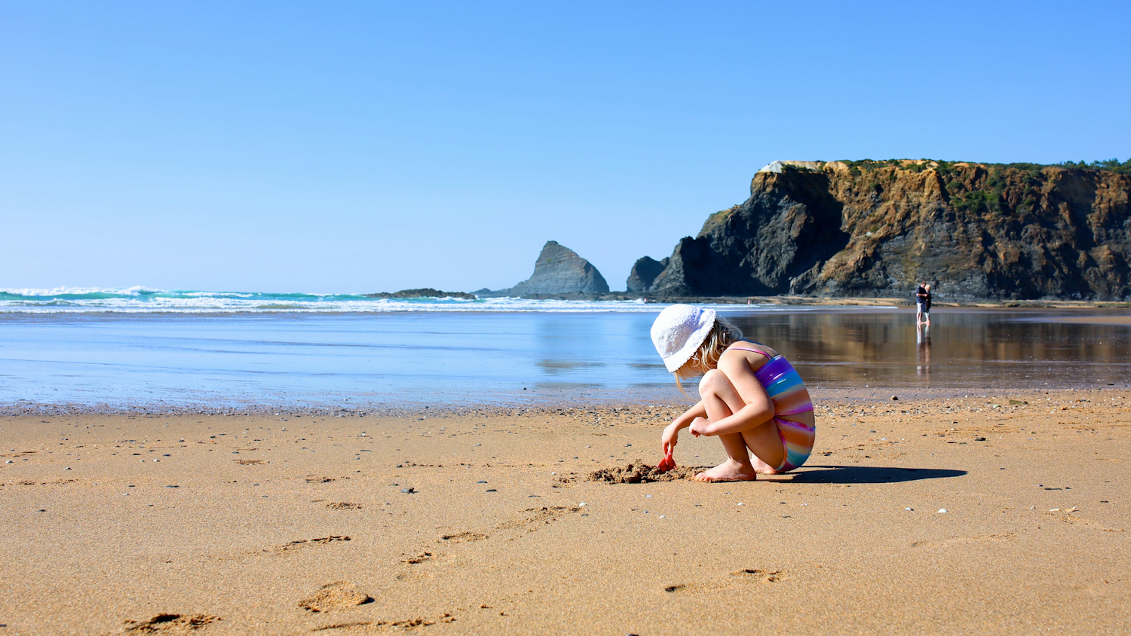 A young child plays with a spade on a sandy beach backed by large cliffs