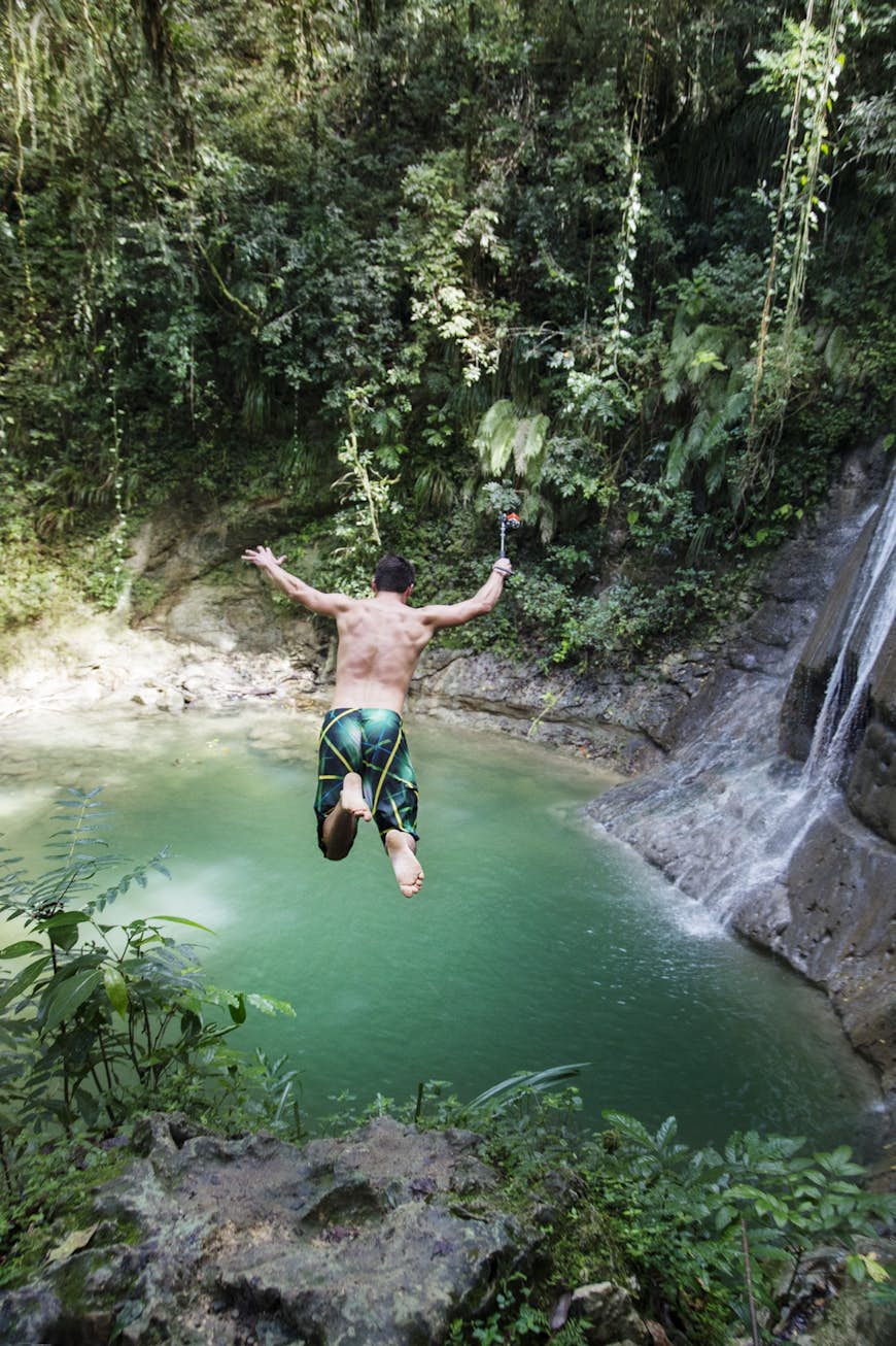 A man jumps off a cliff into a large pool below in a forest in Puerto Rico.   