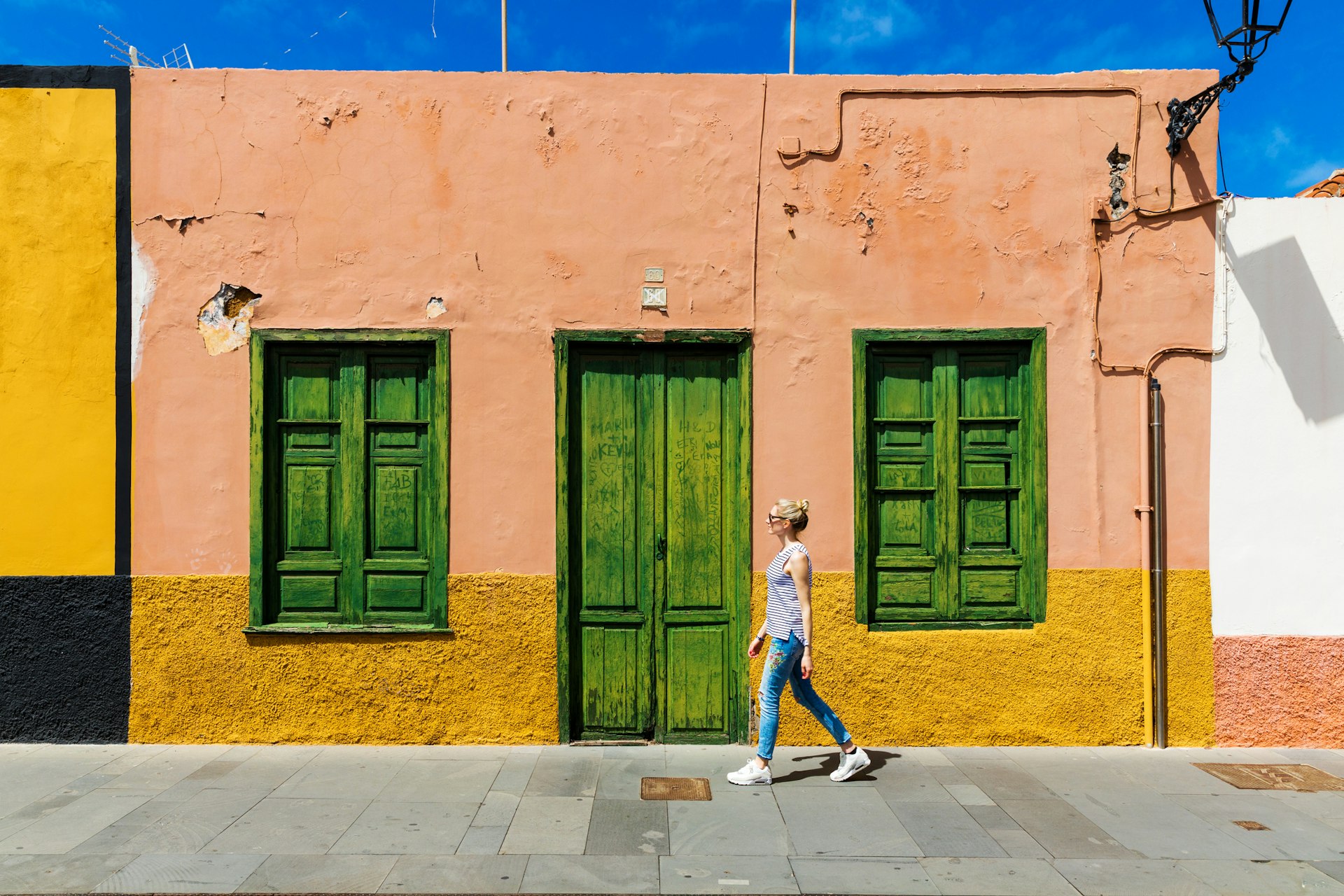 A white woman walks past a single storey pink and yellow house with green shutters in Puerto de la Cruz, Tenerife.