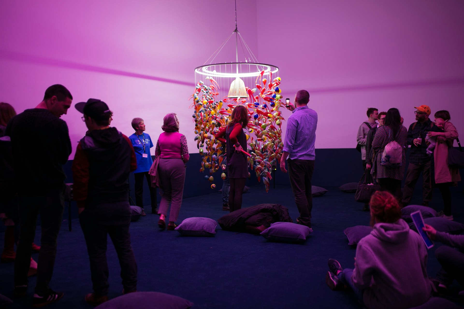 A room of people viewing an art piece that looks like a low chandelier of colored glass