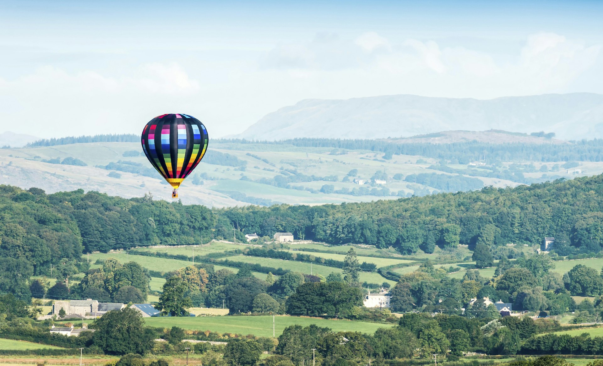 A colorful hot air ballon soars above a green rural scene in Cumbria, England, United Kingdom