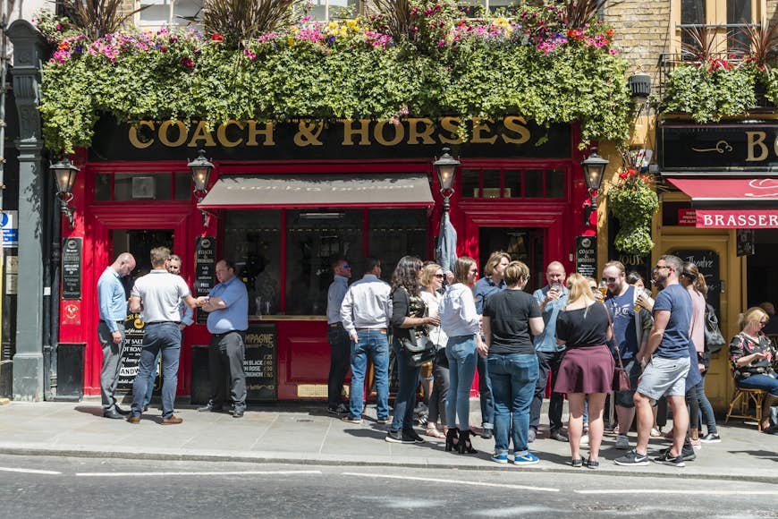 People gather in groups on the sidewalk outside a pub in London enjoying their drinks in the sunshine