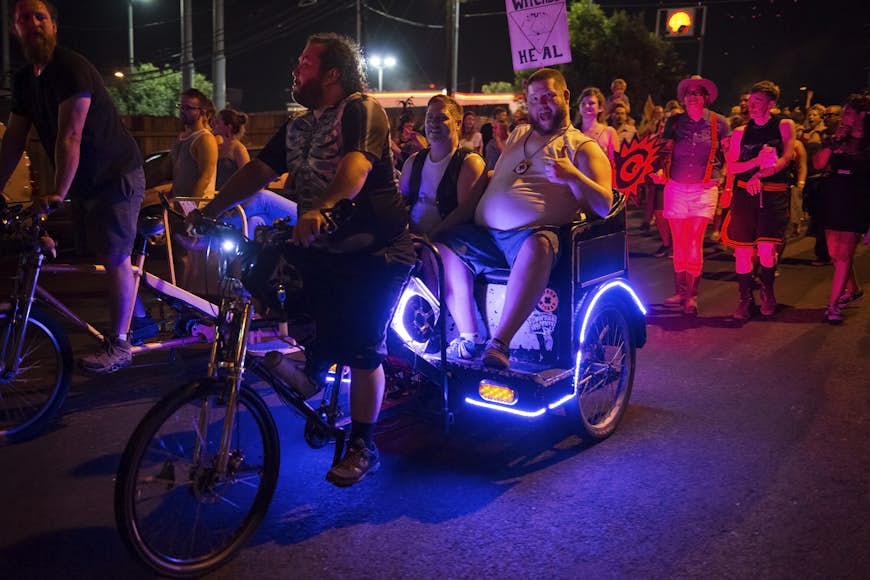 People at a gay parade at night riding a neon-covered tricycle in the city of Austin, Texas