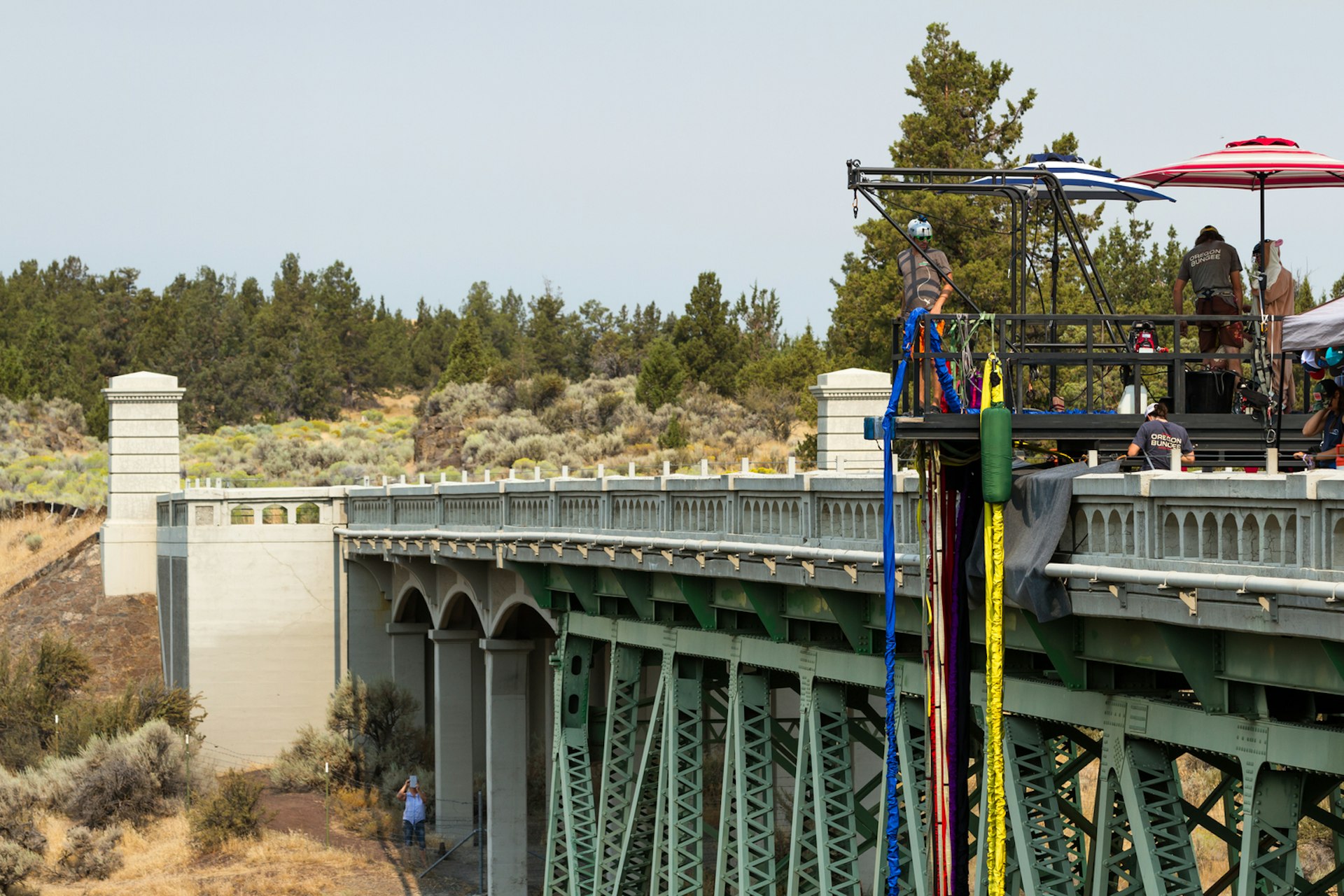A person waits on a bungee jumping platform on a bridge with colorful bungee cords hanging nearby